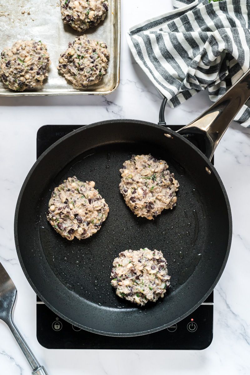 Bean burgers being cooked in a skillet with olive oil and the uncooked patties on the side.